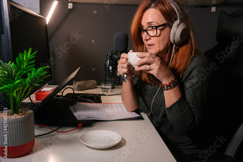 Mujer caucásica de mediana edad tomando un cafe mientras esta grabando un podcast en el despacho de su casa  photo