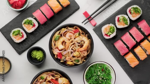 Assorted sushi rolls and wok noodles with shrimps and vegetables in a black bowl, seaweed salad on a white stone background, top view, rotation around its axis. Traditional asian food background