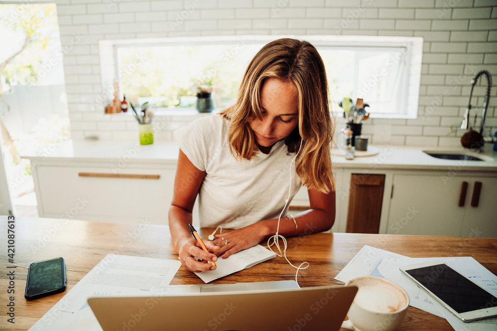 Caucasian female student working from home writing on notepad with laptop listening to music with earphones Caucasian female student working from home writing on notepad with laptop listening to music with earphones