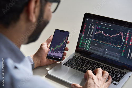 Business man trader investor analyst using mobile phone app analytics for cryptocurrency financial market analysis, trading data index chart graph on smartphone and laptop screen. Over shoulder view