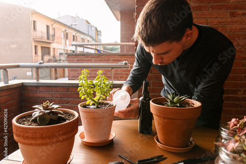 Hombre joven caucásico regando una planta encima de la mesa de la terraza de su casa. Cuidando plantas 