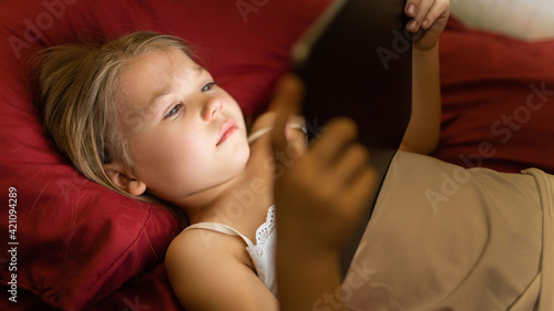 Children and technology concept. A little girl lies in her bed and looks at the tablet and cries.