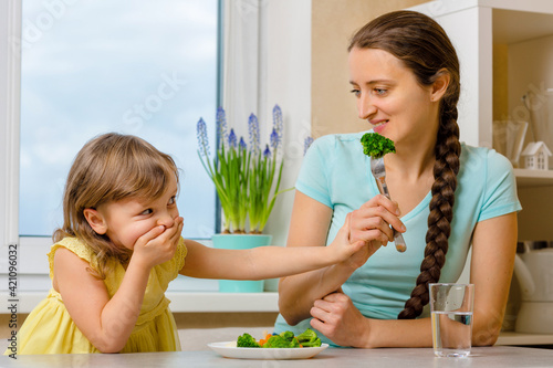 Girl doesn't want eat healthy vegetables. Kid looks with disgust at broccoly. Mother convinces her daughter to eat food. photo