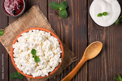 Natural cottage cheese in a traditional clay bowl, next to a wooden spoon, cranberry jam and sour cream in bowls, dark wooden background, top view. Soft curd natural healthy food, wholesome diet food