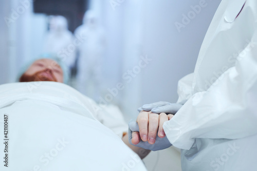Close-up of doctor in protective gloves holding hands of the patient and supporting him while he lying on gurney