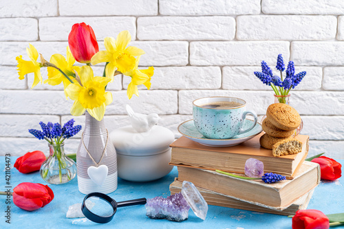 Bouquets of spring flowers, coffee cup, books, cookies, gemstones crystal minerals on blue table on white brick background. Reading and breakfast. Concept spring, gems, hygge and cozy home interior