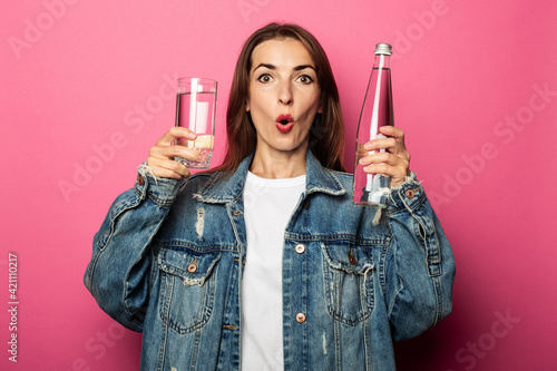 Surprised shocked young woman holding a glass of water and a bottle of water on pink background.