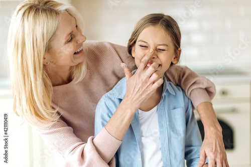 Beautiful caucasian middle aged blonde woman and preschooler girl in the kitchen. Grandma hugging granddaughter, touching her nose finger with flour, they have fun together at home
