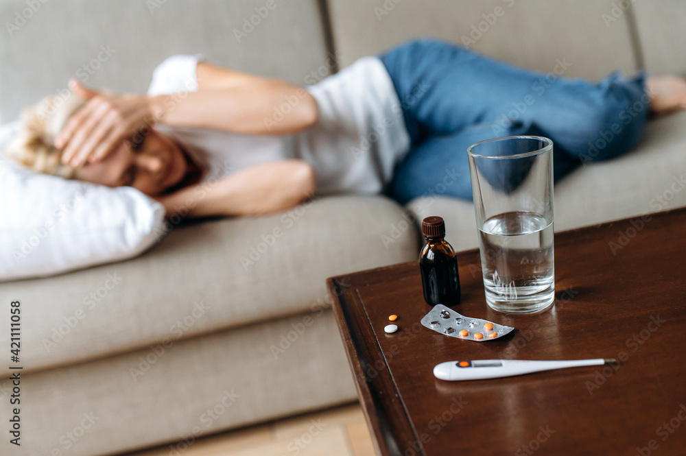 Defocused sick adult woman at home on the couch. A middle-aged caucasian ill woman have headache, clutching her head, a glass of water on the table and a thermometer with pills, need a treatment Defocused sick adult woman at home on the couch. A middle-aged caucasian ill woman have headache, clutching her head, a glass of water on the table and a thermometer with pills, need a treatment