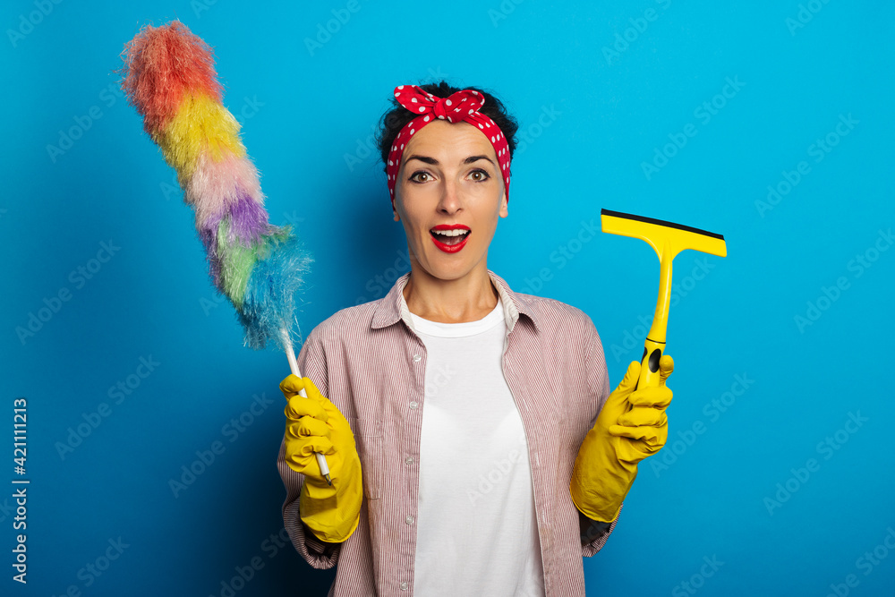 Surprised shocked young woman in gloves holding glass scraper and dust brush on blue background.