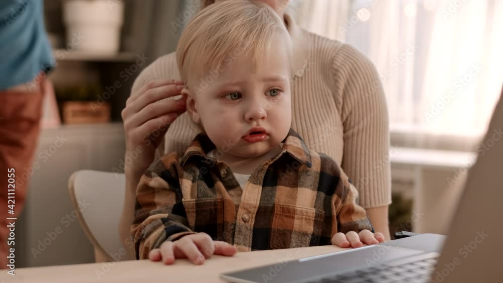 Medium close-up of adorable tear-stained Caucasian toddler wearing plaid shirt, wiping eye with hand, sitting in arms of cropped unrecognizable parent, watching laptop computer Medium close-up of adorable tear-stained Caucasian toddler wearing plaid shirt, wiping eye with hand, sitting in arms of cropped unrecognizable parent, watching laptop computer