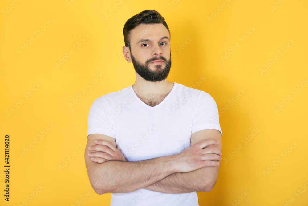 Trendy and stylish serious handsome bearded man in white t-shirt with crossed arms is posing over yellow background Trendy and stylish serious handsome bearded man in white t-shirt with crossed arms is posing over yellow background
