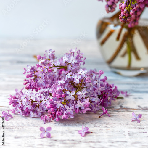 Purple lilac in glass vase on white wooden table. Spring branches of blooming lilac festive bouquet of flowers. Purple lilac branch on table, floral still life on gray wooden background. Square