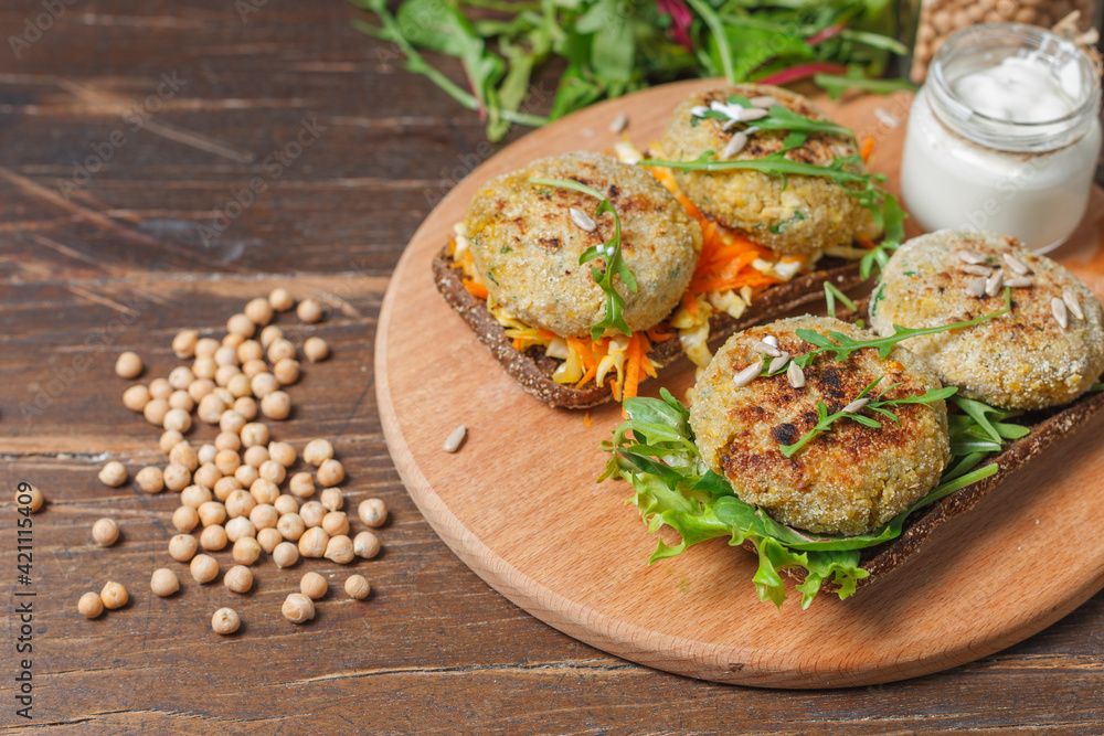 Rye bread sandwiches with chickpea cutlets and vegetable salad. On a wooden board with sauce. And chickpeas on the table.