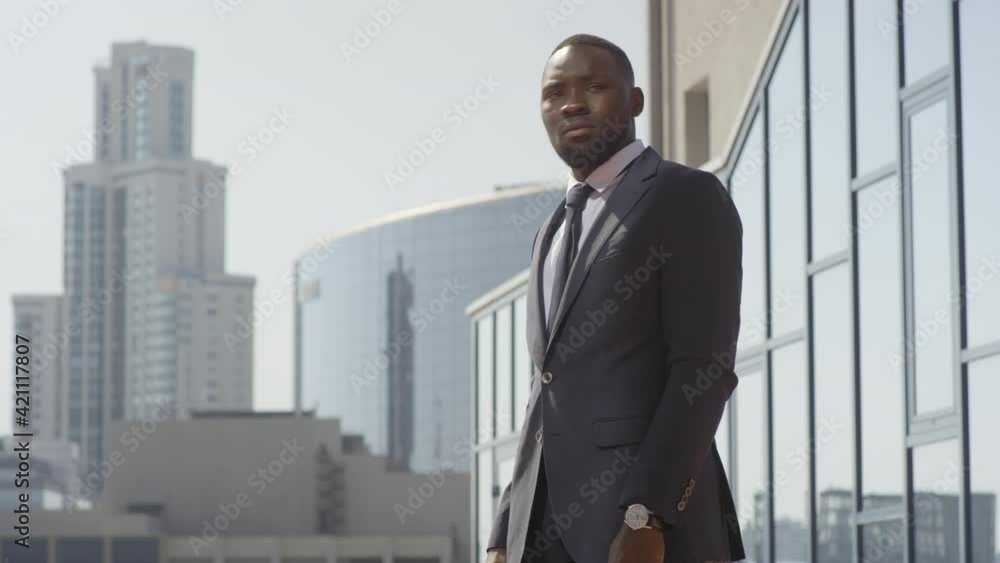 Low-angle medium shot with slowmo of busy african american business man standing outside near modern office waiting for someone looking at his wristwatch being in hurry for business meeting Low-angle medium shot with slowmo of busy african american business man standing outside near modern office waiting for someone looking at his wristwatch being in hurry for business meeting