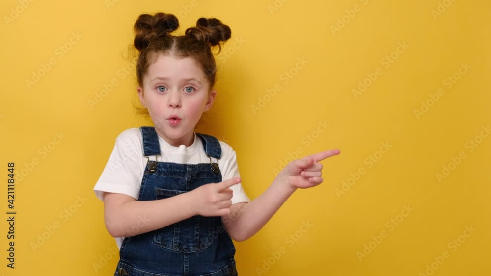 Excited cute little preschool girl pointing back on empty copy space for advertisement text of new store, children goods, services or events, isolated on yellow studio wall. People emotion concept Excited cute little preschool girl pointing back on empty copy space for advertisement text of new store, children goods, services or events, isolated on yellow studio wall. People emotion concept