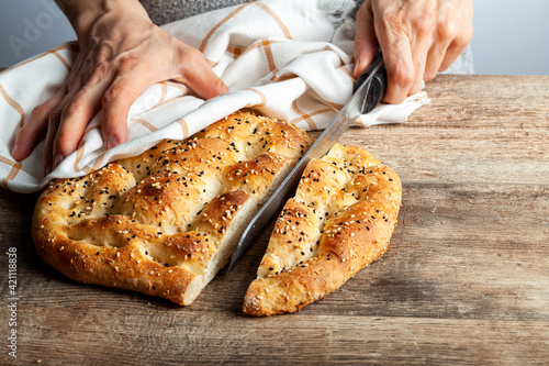 Ramazan pidesi, Turkish flatbread baked during the month of ramadan is a round bread with fennel and sesame seeds on top. It has unique texture and taste. A woman is slicing it on wooden board.
