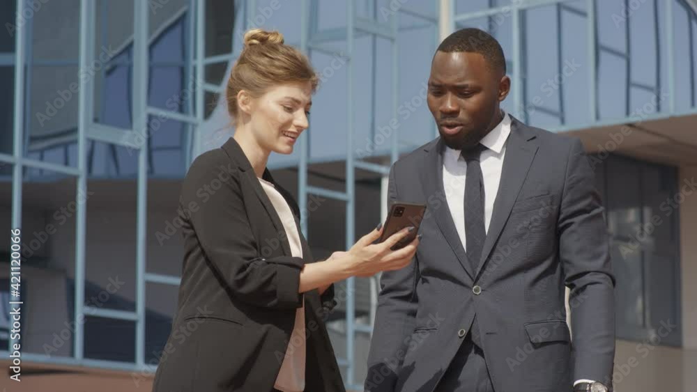 Low-angle medium shot with slowmo of multiethnic business couple looking at smartphone screen together discussing something standing near modern office building on sunny day Low-angle medium shot with slowmo of multiethnic business couple looking at smartphone screen together discussing something standing near modern office building on sunny day
