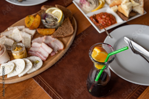 Glass bottle of mulled wine with green plastic drinking straw and slice of orange citrus fruit. Top view table settings with set of appetizers on wooden tray and in white plate