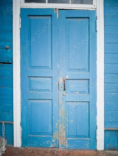 The rickety and curve shabby wooden door to the house. The door is painted with blue paint, the door frame is painted with white paint. © tasslo.studio