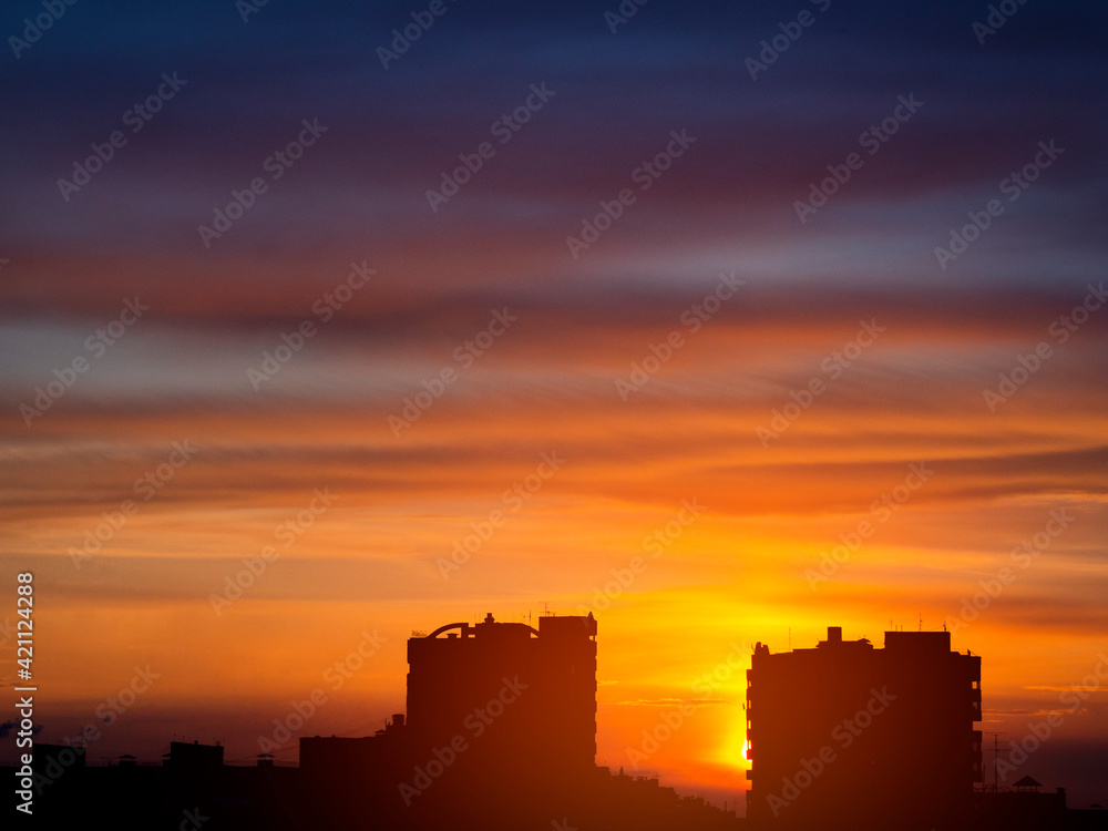 Two dark silhouettes of urban high-rise buildings against the background of an epic spring sunset in purple, red, orange and yellow shades. Glare from the sun, view at the horizon level.
