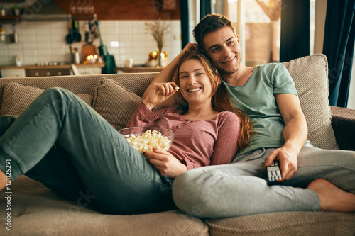 Fotografi Happy couple eating popcorn and watching TV while relaxing on the sofa