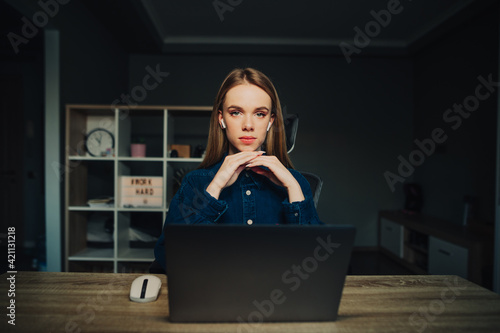 Beautiful woman loyal worker in wireless headphones sitting at home behind a laptop and looking at the camera with a serious face. Freelancer woman working remotely at home.
