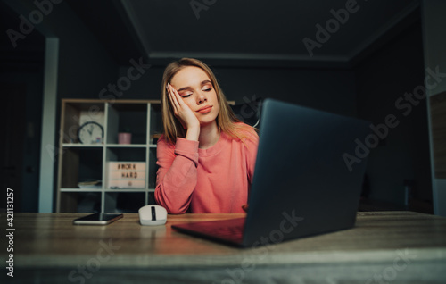 Portrait of upset woman in pink sweater working on laptop at home at work and looking wearily at computer screen with head resting on hand.