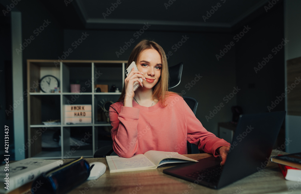 Positive young woman sitting at home at a table with books and a laptop, calling on the phone and looking at the camera with a smile on his face. Positive young woman sitting at home at a table with books and a laptop, calling on the phone and looking at the camera with a smile on his face.
