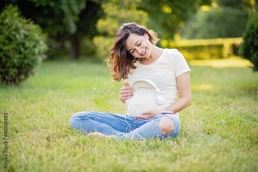Happy young pregnant woman sitting on carpet and listening music on headphones from smartphone at park. Happy young pregnant woman sitting on carpet and listening music on headphones from smartphone at park.