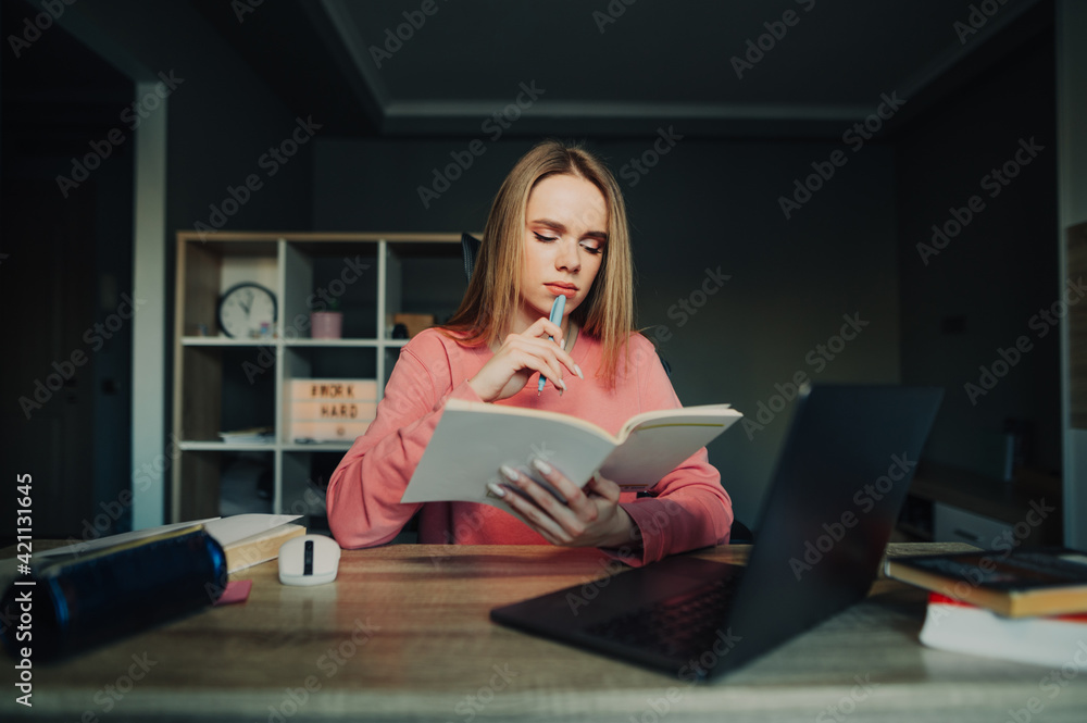 Pensive beautiful student studies at home remotely, sits at a desk and teaches information from a notebook with a pensive face. Woman doing homework. Pensive beautiful student studies at home remotely, sits at a desk and teaches information from a notebook with a pensive face. Woman doing homework.