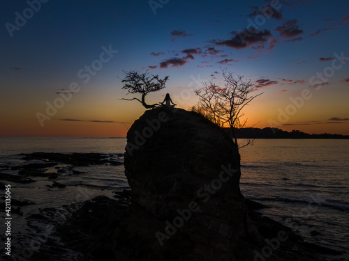 Beautiful and peaceful aerial view of a woman practicing yoga and meditation on a rock over the sunset in a beach 