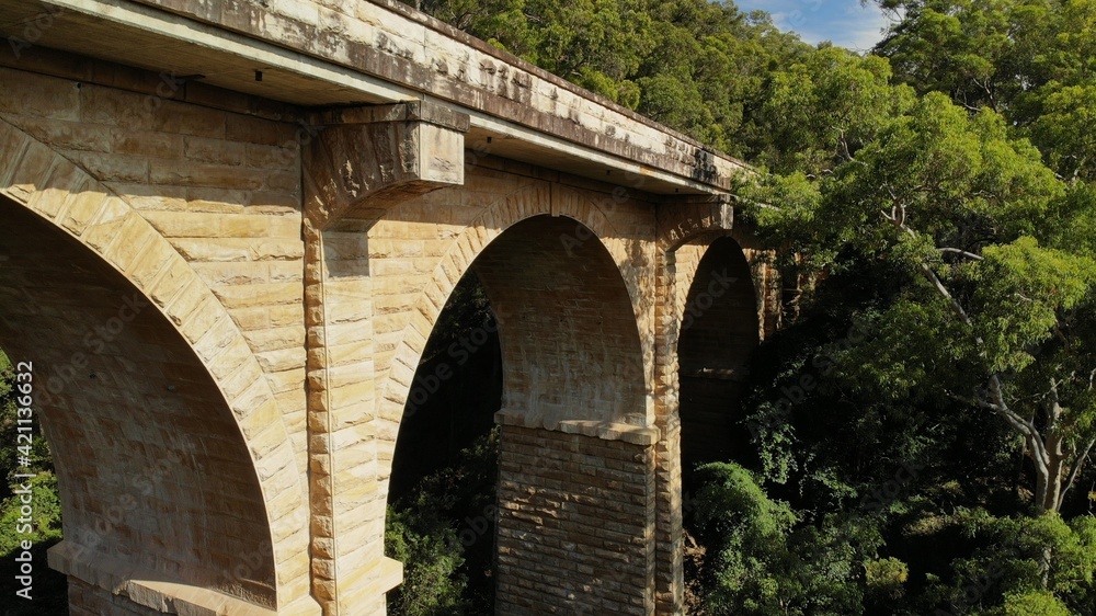 Fototapeta premium Drone images of the Knapsack Bridge, an old sandstone arch bridge, on a sunny spring morning surrounded by green eucalyptus trees.