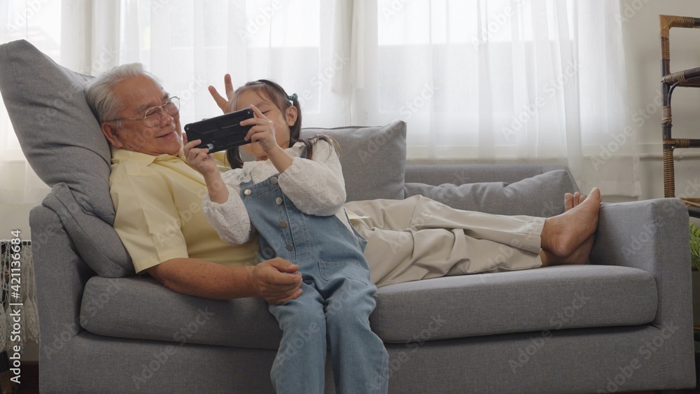 Portrait shot of the happy smiles Asian grandfather and little granddaughter taking selfie photos while sitting on the couch at home. Happy family with spending time together in living room.