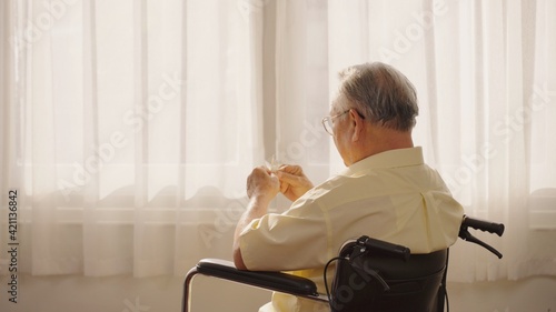 Asian male patient looking outside the window while sitting in wheelchair in living room at retirement home. Asian Thai elderly person sitting lonely in a wheelchair holding a paper bird to play.