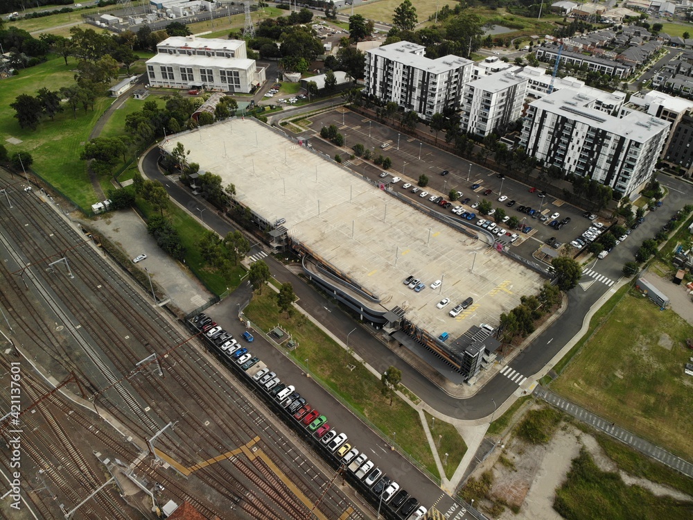 Aerial image of multi storey car park near train line with large apartment blocks in background and green trees.