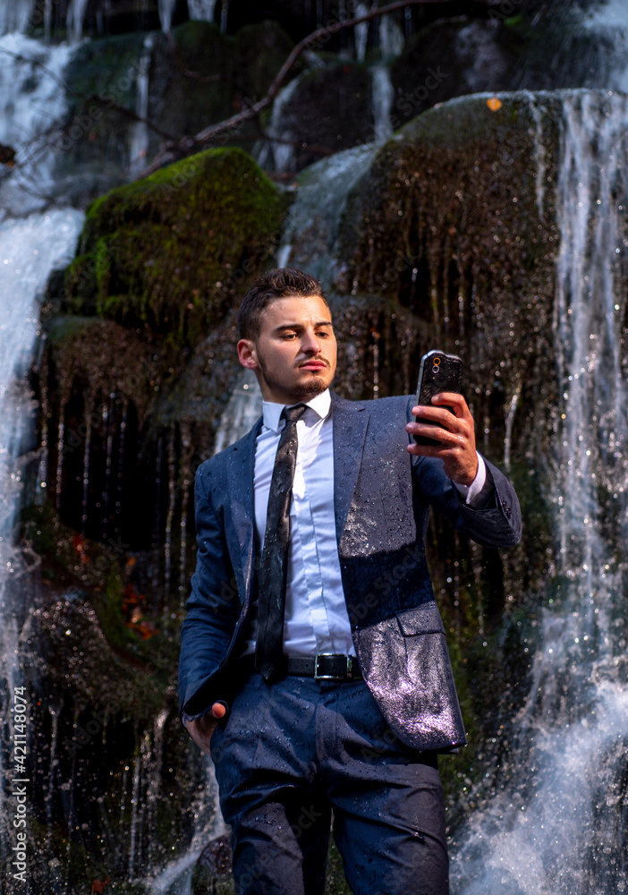 Fototapeta premium Telework employee concept. Handscome business man in suit holding smartphone telephone mobile phone at waterfall background.