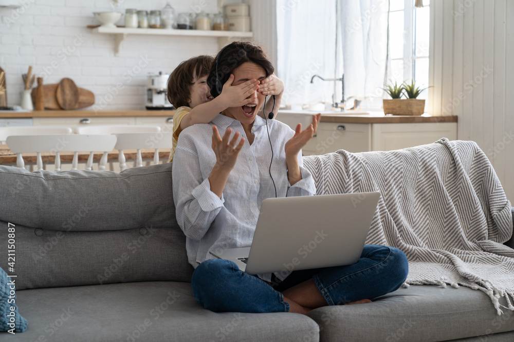 Freelancer woman sitting on couch at home office during lockdown, working on laptop. Playful child distracts from work, covering her mother's eyes, kid making noise and asking attention from busy mom. Freelancer woman sitting on couch at home office during lockdown, working on laptop. Playful child distracts from work, covering her mother's eyes, kid making noise and asking attention from busy mom.