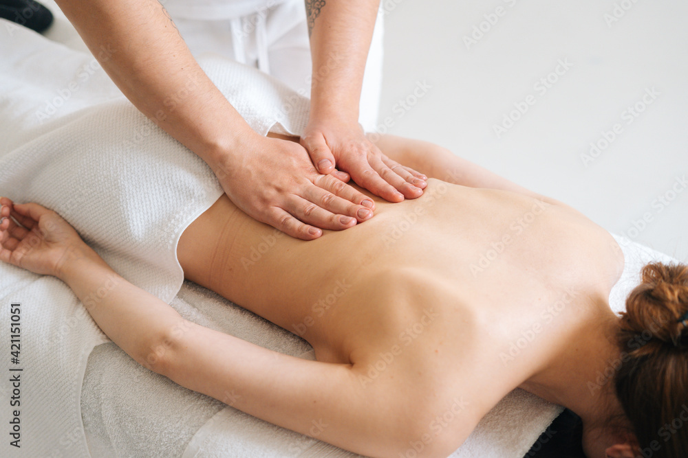Top view of male masseur massaging lower back of young woman lying on massage table at light spa salon. Experienced chiropractor performs wellness treatments for lady with back pain.