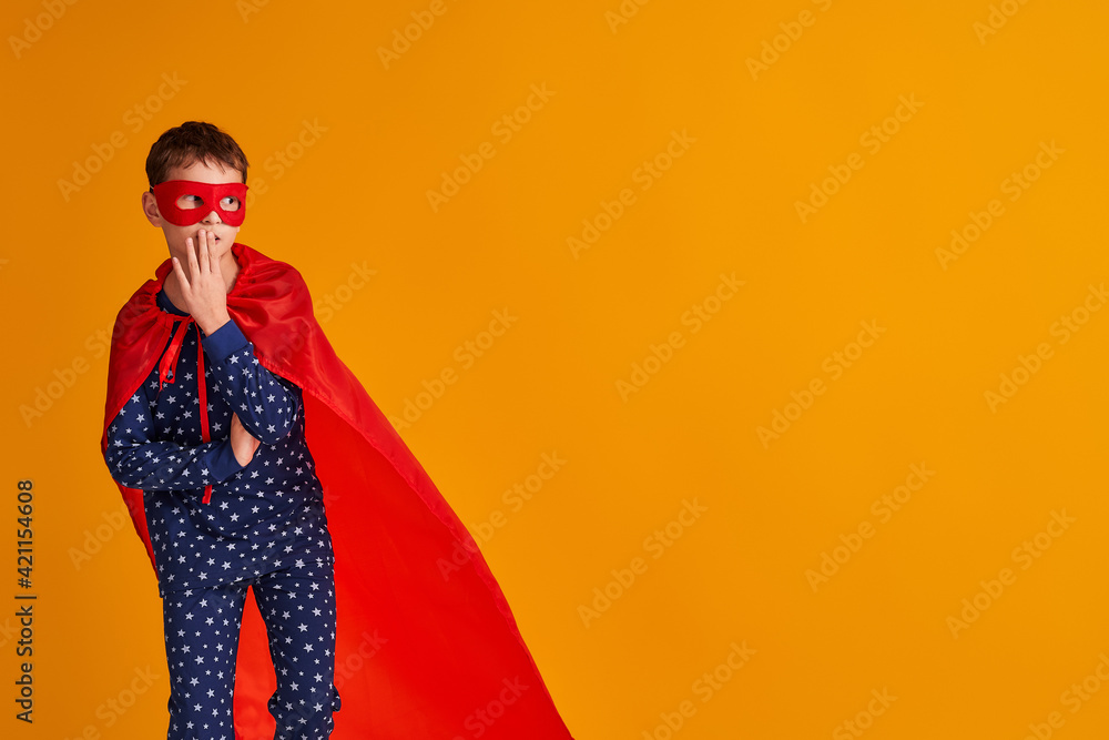 surprised child is playing in a superhero costume. the boy is dressed in a red cloak and mask, holding his hand to his mouth and looking doubtfully to the side on a yellow background. Copy space surprised child is playing in a superhero costume. the boy is dressed in a red cloak and mask, holding his hand to his mouth and looking doubtfully to the side on a yellow background. Copy space