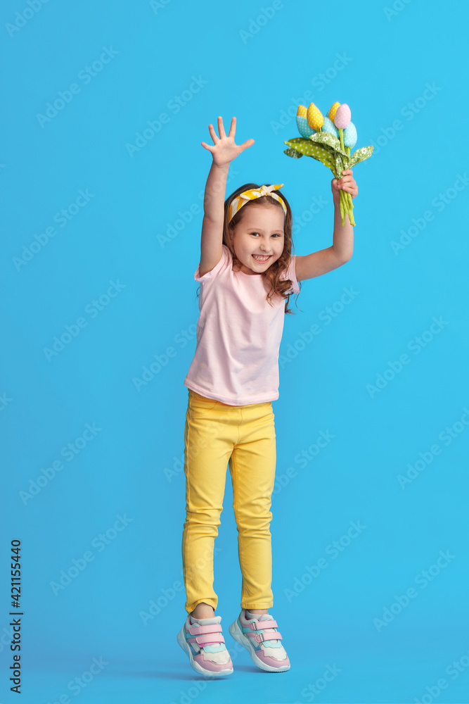 charming little girl with curly hair and headband with bouquet colorful tulips made fabric jumps to her full height on blue background studio. child in fashionable summer clothes. Dynamic image