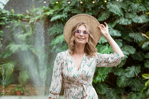 beautiful young blonde woman in a straw hat and glasses posing in the park against a background of greenery