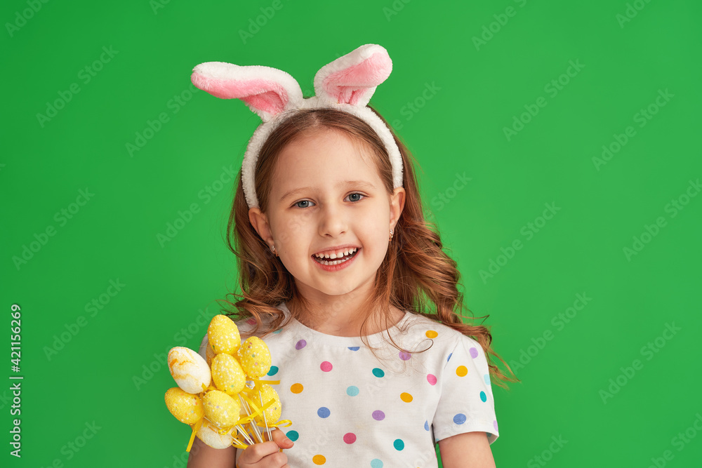 Portrait of a cute little girl wearing the ears of an Easter bunny holding colorful eggs standing on a green background on Easter day. The child is happy and celebrates Easter Portrait of a cute little girl wearing the ears of an Easter bunny holding colorful eggs standing on a green background on Easter day. The child is happy and celebrates Easter