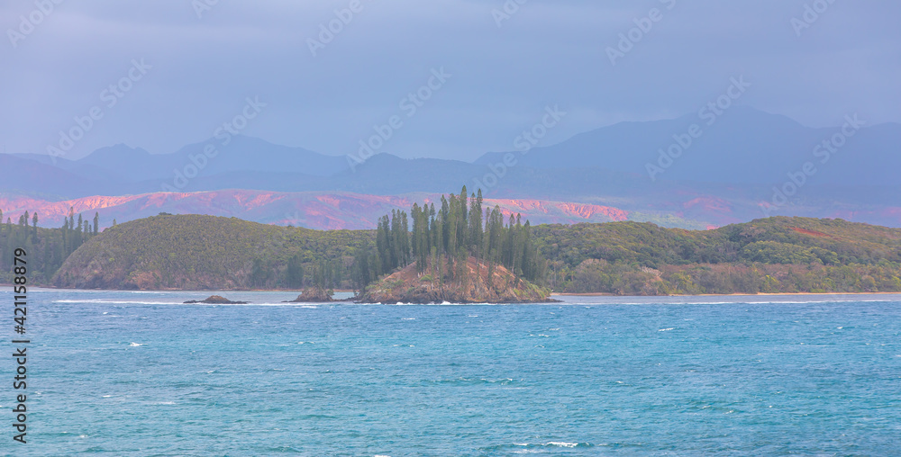 Beautiful small island covered by pine trees forms the central focus for this view of the coastline with colourful highlights from a small break in the clouds on a very hazy day, Noumea,New Caledonia