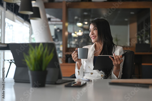 Thoughtfully businesswoman in earphone relaxing in office holding cup of coffee looking away.