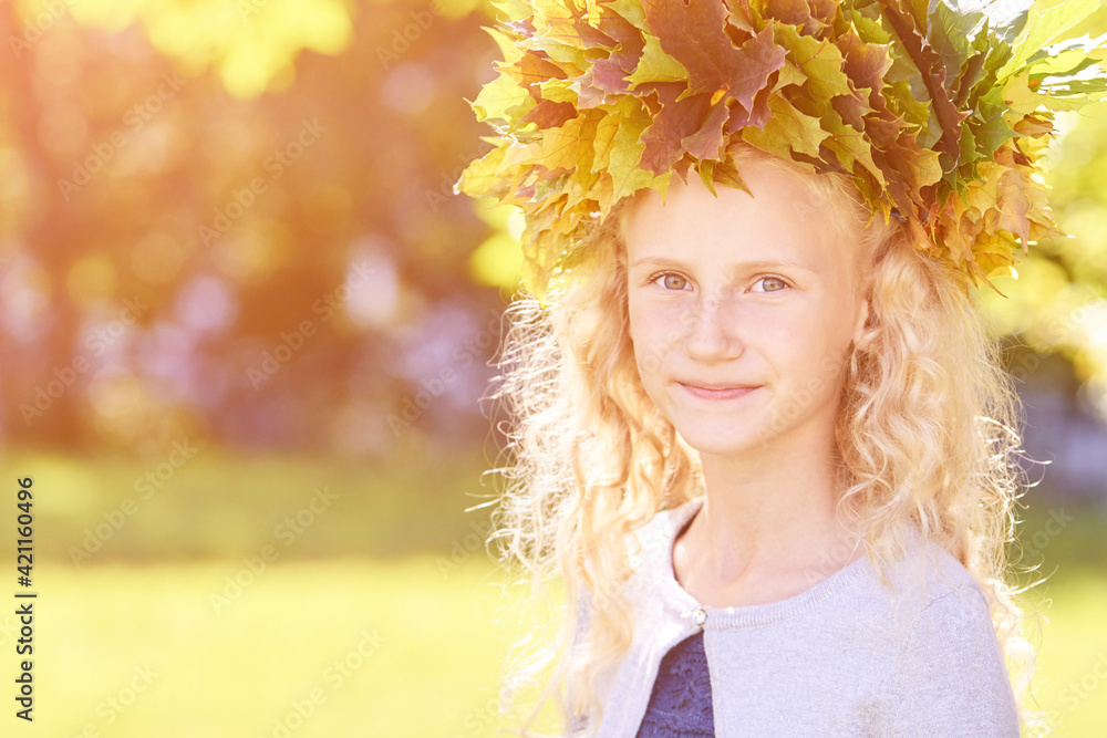 Fototapeta premium Little girl with autumn maple wreath. Yellow and green colors. Backyard nature. Female child portrait. Happy people. Model posing at park