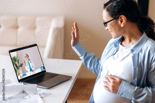 Online medical consultation. Mixed race adult pregnant woman talks with a female doctor by video call uses laptop, getting medical consultation, receives answers to questions and recommendations