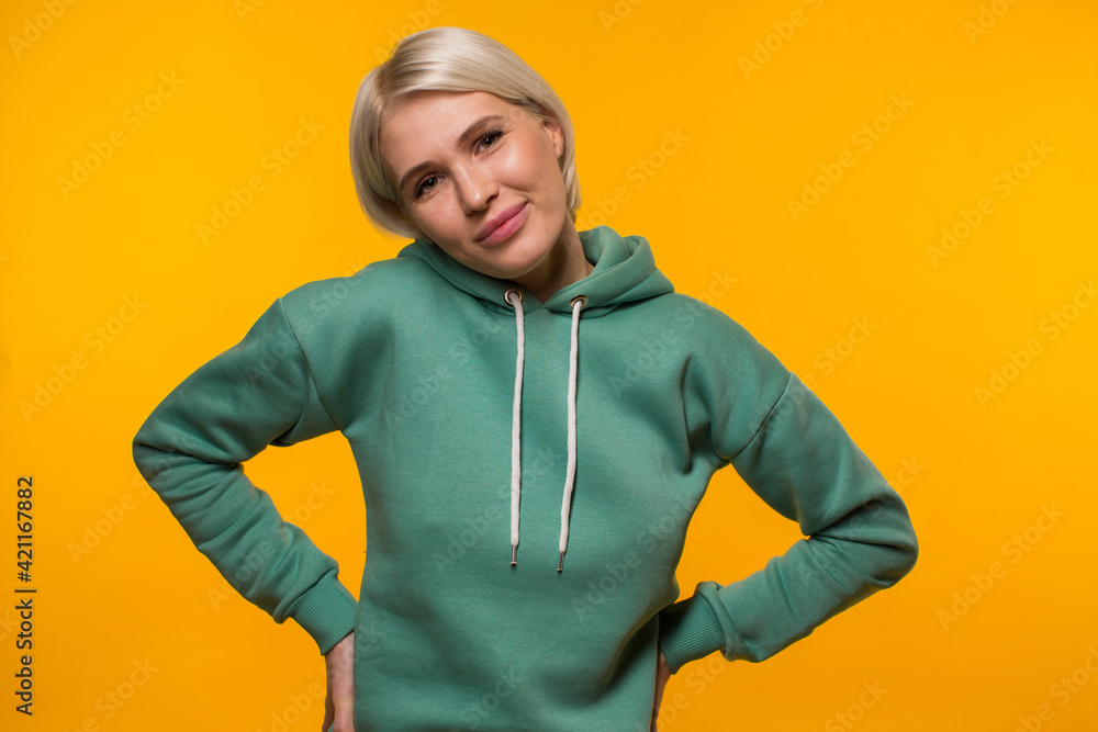 Portrait of a female student in a dark green hoodie who stands in the studio on a bright yellow background smiles and looks directly at the camera. Bright photo. Model shooting. High quality photo Portrait of a female student in a dark green hoodie who stands in the studio on a bright yellow background smiles and looks directly at the camera. Bright photo. Model shooting. High quality photo
