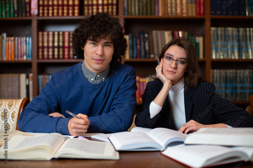 photo of the educational process in the college library. Two students sit near a set of books and look directly at the camera. High quality photo photo of the educational process in the college library. Two students sit near a set of books and look directly at the camera. High quality photo