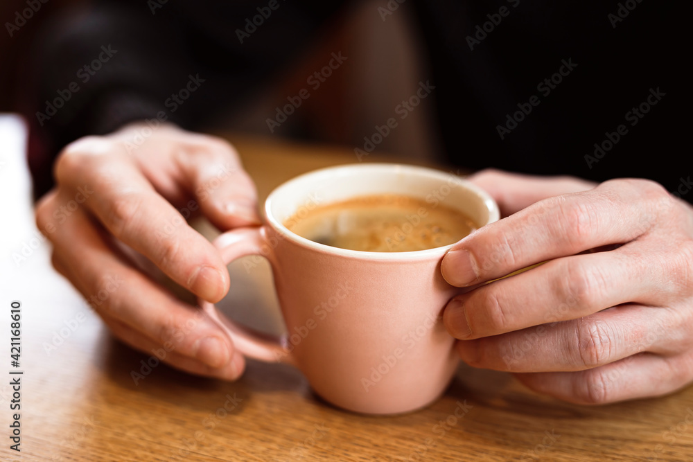 Man holding a pink ceramic cup of hot black coffee in his hands, sitting at the wooden table in a cafe. Close-up photo Man holding a pink ceramic cup of hot black coffee in his hands, sitting at the wooden table in a cafe. Close-up photo